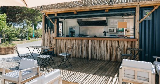 Comptoir de restauration habillé de bois, ouvert sur une terrasse avec tables et fauteuils sous une pergola.