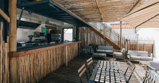 Comptoir de restauration habillé de bois, ouvert sur une terrasse avec tables et fauteuils sous une pergola.