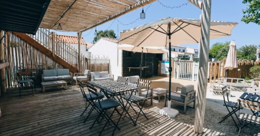 Tables, chaises et fauteuils sur une terrasse en bois ombragée par une pergola et des parasols.