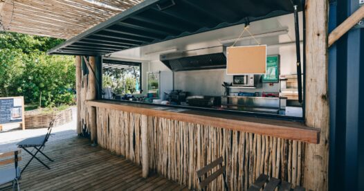 Comptoir de restauration habillé de bois, ouvert sur une terrasse avec tables et fauteuils sous une pergola.