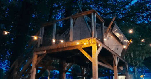 Tente bivouac sur pilotis éclairée par une guirlande à la tombée de la nuit, avec une table de pique-nique en dessous.