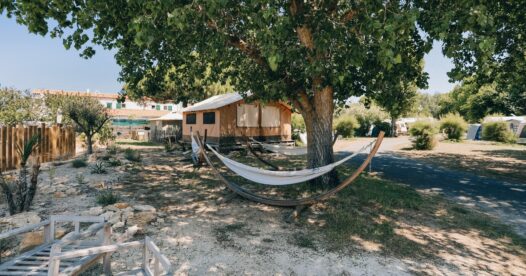 Hamac sur un support en bois à l’ombre d’un grand arbre, près des hébergements du camping.