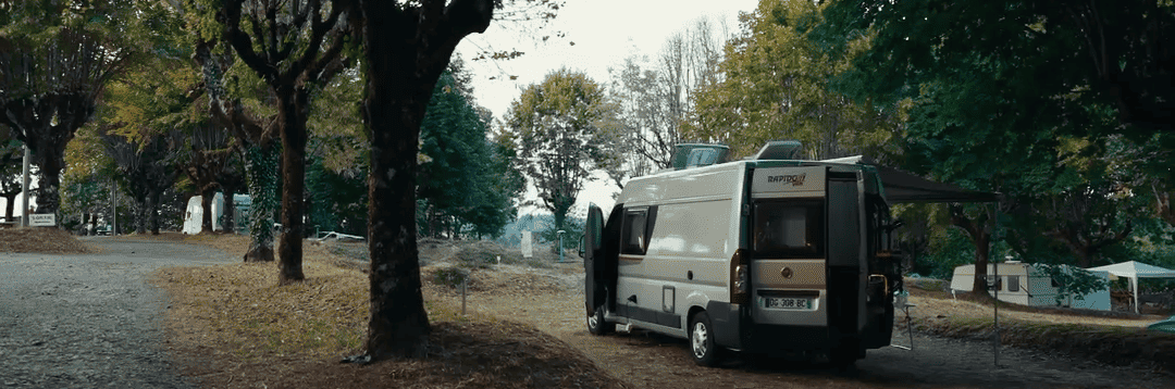 Fourgon aménagé stationné sur un emplacement de camping sous de grands arbres.