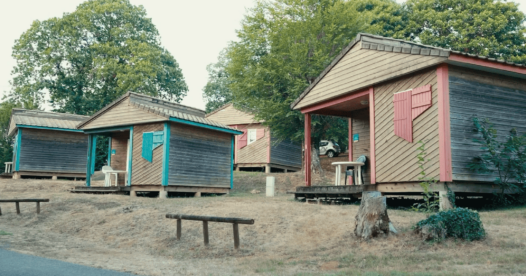 Chalets en bois aux volets colorés, avec terrasses couvertes, le long d’une allée du camping.