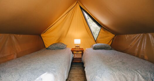 Deux lits simples disposés sous la toile d’une tente, avec une lampe de chevet entre les couchages.