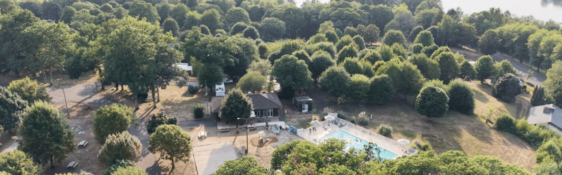 Vue aérienne du camping arboré et de sa piscine extérieure, avec un lac en arrière-plan.