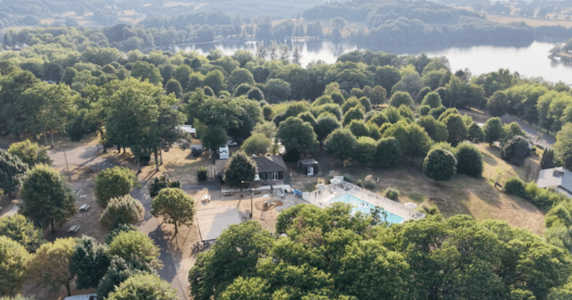 Vue aérienne du camping arboré et de sa piscine extérieure, avec un lac en arrière-plan.