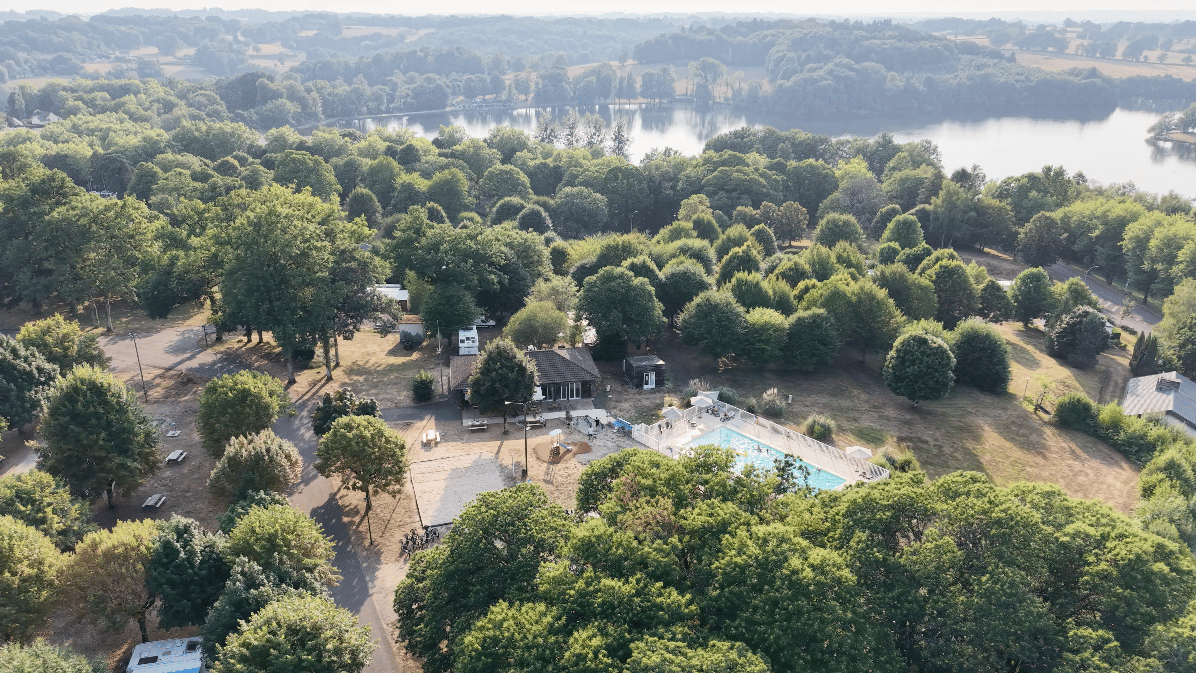 Vue aérienne du camping arboré et de sa piscine extérieure, avec un lac en arrière-plan.