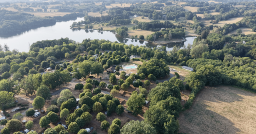 Vue aérienne du camping de Bournazel et de sa piscine, entourés d’arbres, avec un lac en arrière-plan.