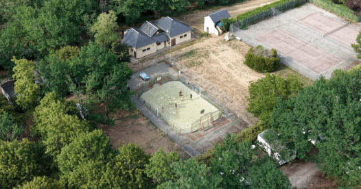 Vue aérienne de deux courts de tennis et d’un terrain multisports entourés d’arbres.
