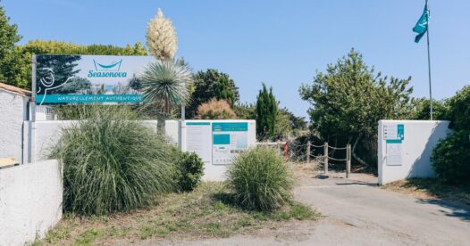 Entrée du camping avec panneau Seasonova, drapeaux turquoise et massifs de végétation.