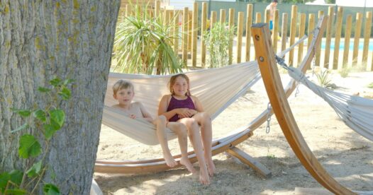 Deux enfants assis dans un hamac en toile suspendu à un support en bois, à l’ombre d’un arbre.