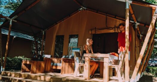 Vacanciers sur la terrasse couverte d’une tente lodge, autour d’une table en bois sous les arbres.