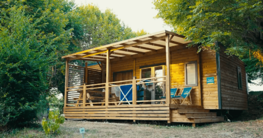 Mobil-home avec bardage en bois et terrasse couverte équipée de chaises, entouré d’arbres.