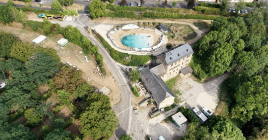 Vue aérienne d’une piscine extérieure et d’un grand bâtiment en pierre entourés d’arbres.