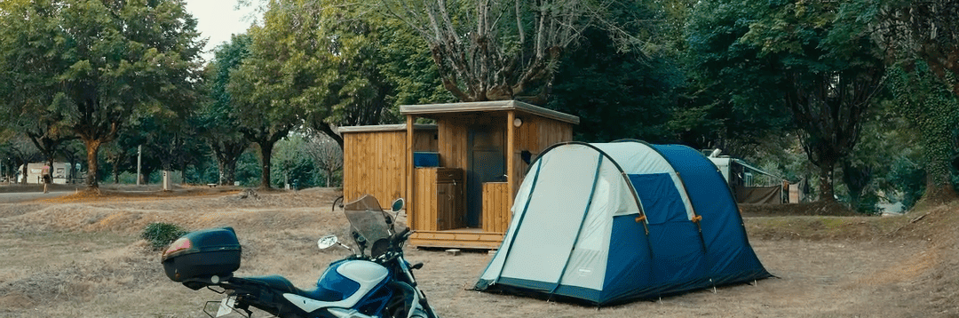 Moto stationnée à côté d’une tente bleue sur un emplacement de camping arboré.