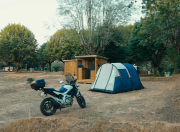 Moto stationnée à côté d’une tente bleue sur un emplacement de camping arboré.