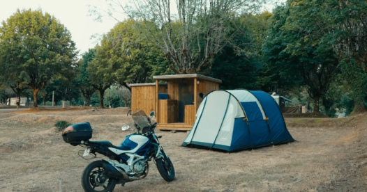 Moto stationnée à côté d’une tente bleue sur un emplacement de camping arboré.