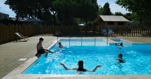 Plusieurs vacanciers se baignent dans la piscine extérieure du camping, entourée d’une clôture en bois.