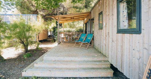 Terrasse en bois avec quelques marches, deux chiliennes turquoise et une table sous l’auvent.