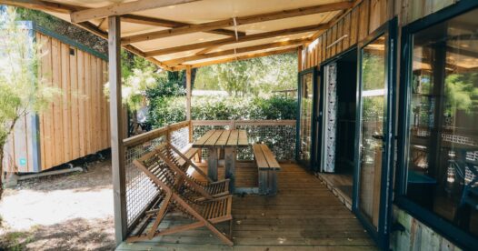 Table avec bancs et chaises en bois sous le toit d’une terrasse, le long de la baie vitrée de l’hébergement.